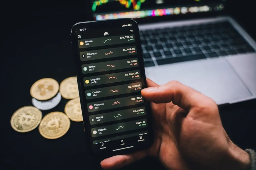 A person using a cryptocurrency trading app on a smartphone with Bitcoin coins and a laptop in the background, representing digital investment tools for wealth-building.